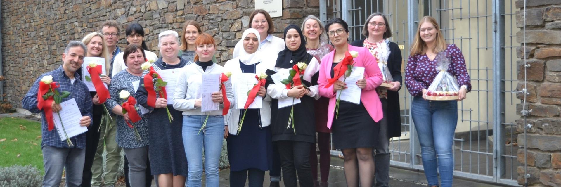 Glücklich halten die Absolventinnen und Absolventen ihre Zertifikate und Rosen in den Händen. Mit dem Abschluss im Bereich qualifizierende Vorbereitung zu einer Ausbildung oder Beschäftigung in der  Alten- & Krankenpflege finden sie ihren beruflichen Weg.